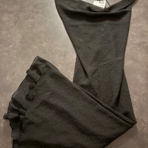 Svart glittrig långklänning med smala band - Supersnygg svart långklänning med smala axelband och glittriga detaljer över hela tyget. Klänningen har en lätt draperad urringning och volangdetalj nertill. Perfekt för dig som vill sticka ut med en elegant och trendig look.