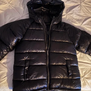 Svart pufferjacka Supply & Demand - Svart pufferjacka från Supply & Demand med glansig finish, huva och broderad logga på ryggen. Jackan har två fickor fram, dragkedja och patch på ärmen. Perfekt för kalla dagar med sin tjocka vaddering och stilrena look.STORLEK 10-12 år.