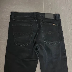 Nudie jeans  - Svarta jeans från Nudie Jeans med klassisk femficksdesign och broderade bakfickor. Jeansen har smal passform och raka ben, tillverkade i ett stretchigt bomullsmaterial för extra komfort. Perfekta för en stilren och enkel look.