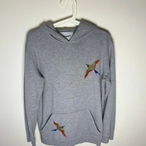  Axel Arigato hoddie  - Grå hoodie från Axel Arigato med broderade färgglada fåglar på bröstet och fickan. Tröjan har huva, lång ärm och känguruficka framtill. Perfekt för en chill och stilren look. Hoddien är i storlek xs men sitter lite längre så mer som en S, Frågor/funderingar hör av dig 🤙🏽 