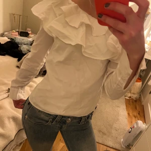 Vit blus med volang från H&M - Säljer en vit blus från H&M med bred volang längs den offshoulder skurna halsen. Blusen har lång ärm med manschett och är gjord i ett lätt bomullsmaterial. Den är i ”skjorttyg”. Köpt secondhand men i bra skick! Jätte söt 💕