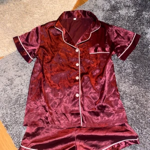  satinshorts med vit kant - Snygga vinröda shorts i satin med vit kant längs benen. De har en mjuk och glansig yta som känns lyxig och chill, perfekta för att hänga hemma eller sova i. Matchar med en kortärmad skjorta i samma stil för en komplett pyjamaslook. På bilden ser dem röda ut men dem är egentligen lite mer åt de röd/lila hållet 