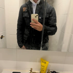 Svart quiltad Barbour jacka - Quiltad svart jacka från Barbour med klassisk design och snygga detaljer som tryckknappar, dragkedja och patch på bröstet. Jackan har lång ärm, ståkrage och Barbour-logga på fickan. Perfekt för dig som gillar stilren och tidlös look.