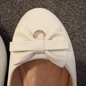 Ballerina skor  - Vita ballerinaskor med rundad tå. Skorna har en dekorativ rosett med en strassdetalj i mitten. Aldrig använt eftersom den är för stor för mig 