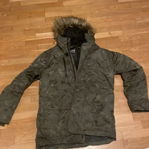 Camouflage parkajacka från Helly Hansen - Parkajacka från Helly Hansen i mörkgrön camouflage med fuskpäls på huvan. Jackan har flera fickor, dragkedja och knäppning framtill. Perfekt för kalla dagar med sin långa modell och fodrade insida. Snygg och funktionell design för dig som gillar streetstyle. Storleken var fel men tyvärr togs prislappen bort.