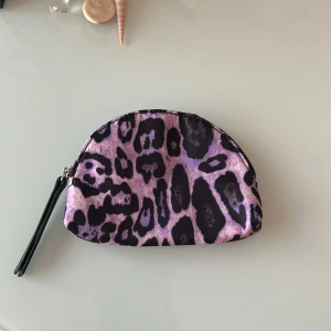 Lila leopardmönstrad plånbok - Cool plånbok i halvmåneform med lila och svart leopardmönster. Väskan har dragkedja och en svart handledsrem. Perfekt för att förvara småsaker eller smink. Materialet är mjukt tyg och insidan är svart.