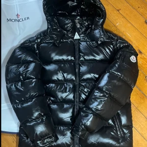 Svart Moncler pufferjacka med huva - Säljer en svart pufferjacka från Moncler med glansig finish och huva. Jackan har Moncler-logga på ärmen, dragkedja framtill och ser riktigt fet ut för vintern. Insidan har en cool serietidningspatch. Perfekt för dig som vill ha en stilren och varm jacka.