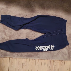 Mörkblå mjukisbyxor från Hollister - Mörkblå mjukisbyxor från Hollister med vit broderad logga längs ena benet. Byxorna har resår och snörning i midjan samt muddar vid bensluten. Tillverkade i mjuk bomull för maximal komfort.