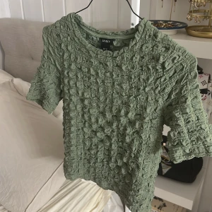 Grön strukturerad topp från Lindex - Säljer en ljusgrön topp från Lindex i storlek XS. Toppen har korta ärmar och är gjord i ett strukturerat, bubbligt material som ger en cool och unik look. Perfekt för dig som vill sticka ut med en trendig och bekväm stil.