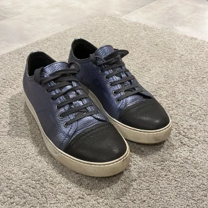 Lanvin blå och svarta sneakers i skinn - Snygga sneakers från Lanvin i blått och svart skinn med vit sula. Skorna har rund tå, svarta skosnören och en klassisk låg profil. Kommer med original dustbag från Lanvin. Perfekta för dig som gillar stilrena och exklusiva sneakers.