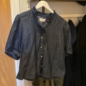 Mörkblå jeans topp med volang  - Säljer en mörkblå blus från Holly & Whyte by Lindex i storlek M. Blusen har korta puffärmar, knappar framtill och en söt volangdetalj vid halsen. Materialet är mjukt och känns som bomull, perfekt för dig som gillar en chill och snygg look.