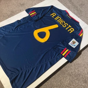 Spaniens matchtröja Iniesta VM 2010 Adidas - Säljer en officiell Spanien fotbollströja från VM 2010 med Iniesta och nummer 6 på ryggen. Tröjan är marinblå med gula och röda detaljer, broderat landslagsemblem och Adidas-logga. Korta ärmar med klassiska ränder och Climacool-material för bästa komfort.