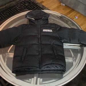 Svart pufferjacka från Hoodrich - Svart pufferjacka från Hoodrich med huva och vit logga på bröstet samt insidan av huvan. Jackan har dragkedja framtill, två sidofickor och quiltad design. Perfekt för kalla dagar med sin tjocka och fluffiga look, lite förstörd i högra sidan av jackan men att sy kommer enkelt fixa. 155cm 