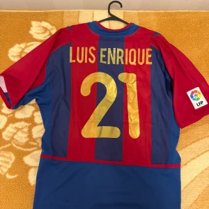 Barcelona Luis Enrique 21 Nike tröja - Klassisk FC Barcelona matchtröja med Luis Enrique och nummer 21 på ryggen. Rödblåa vertikala ränder, officiellt klubbmärke och Nike-logga framtill. Kortärmad modell i lätt polyester, LFP-patch på ärmen. Perfekt för samlare eller fans av spansk fotboll.