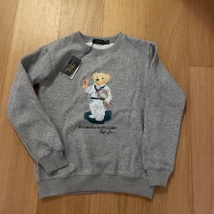Grå sweatshirt Polo Ralph Lauren  - Grå sweatshirt från Polo Ralph Lauren med ikoniska Polo Bear-trycket på framsidan där nallen är klädd i tennisoutfit. Tröjan har rund hals, ribbade muddar och är långärmad. Perfekt för dig som gillar sportiga och snygga detaljer.