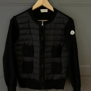 Svart cardigan från Moncler - Svart pufferjacka från Moncler med quiltad front och stickade ärmar. Jackan har rund hals, dragkedja framtill och Moncler-logga på vänster ärm. Materialmix av dun och stickad polyester ger en schysst kontrast. Perfekt för dig som gillar stilren design och premiumkänsla.