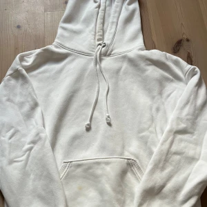 Vit hoodie med snörning och ficka - Vit hoodie med klassisk huva och snörning, samt en stor magficka framtill. Tröjan har långa ärmar och är gjord i mjukt bomullsmaterial. Perfekt för chill dagar och enkel att matcha med jeans eller mjukisbyxor. den har tyvärr några fläckar på sig, men de är inte som märkvärdiga