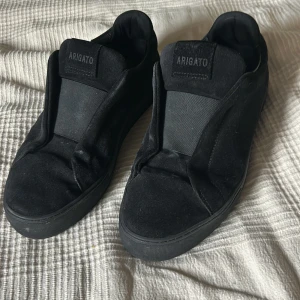 Svarta sneakers från Axel Arigato - Säljer ett par helsvarta sneakers från Axel Arigato i mocka med minimalistisk design. Skorna har elastisk panel över vristen istället för klassisk snörning och en platt sula. Perfekta för dig som gillar stilrena och cleana skor.de ingår även lådan och ett par extra skosulor ca som är bra att ha om dem är lite för stora för att fylla utrymme 