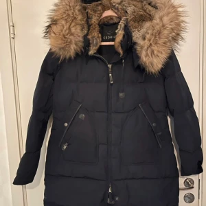 Mörkblå dunjacka med päls från Cedrico - Säljer en mörkblå dunjacka från Cedrico i storlek L. Jackan har en stor fluffig huva med päls, praktiska fickor med knappar och dragkedja framtill. Den är quiltad och har snörning nedtill för extra passform. Perfekt för kalla vinterdagar.