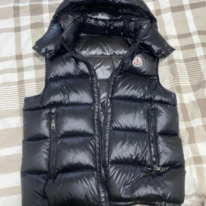 Svart dunväst från Moncler - Snygg svart dunväst från Moncler Storlek M med glansig finish, hög krage och avtagbar huva. Västen har två dragkedjeförsedda fickor framtill och klassisk Moncler-logga på bröstet. Perfekt för lager-på-lager och streetstyle.