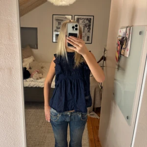 Mörkblå ärmlös blus med volang - Supersöt mörkblå blus med volangdetaljer längs axlarna. Den är ärmlös och har en avslappnad, lite lös passform. 