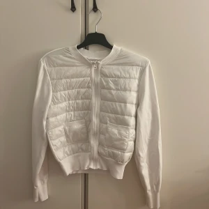 Vit quiltad pufferjacka Gina Tricot - Snygg vit pufferjacka från Gina Tricot med quiltad framsida och två praktiska fickor. Jackan har ribbad krage och muddar, samt dragkedja framtill. Perfekt för dig som gillar stilren och clean look.