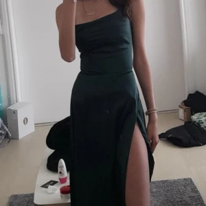 Mörkgrön  långklänning - Snygg mörkgrön långklänning med design och hög slits på sidan. Klänningen har en elegant och stilren look, perfekt för dig som vill sticka ut. Materialet är mjukt och har en lätt glansig finish som ger en lyxig känsla.