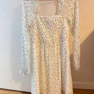 Vit blommig långärmad klänning - Söt vit klänning med små gröna blommor och smockad överdel. Långa, transparenta ärmar med volangdetaljer vid axlarna ger en romantisk vibe. Perfekt för dig som gillar en feminin och fräsch stil. Lätt och luftig, passar bra till varmare dagar.