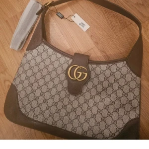 Axelväska och plånbok GG monogram - Säljer en snygg axelväska och matchande plånbok från med klassiskt GG-monogram i beige och brunt. Väskan har guldiga GG-detaljer, bred axelrem och insida med fack och dragkedja. Plånboken har dragkedja och samma ikoniska mönster och färgkombination.