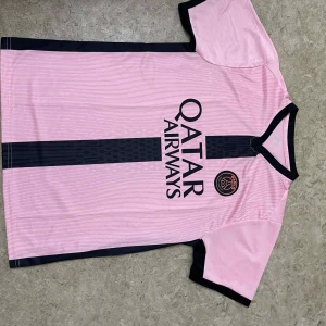 Rosa PSG fotbollströja Qatar Airways - Snygg rosa PSG fotbollströja med svart vertikal rand och svarta detaljer vid krage och ärmslut. Tryck med Qatar Airways på framsidan och PSG-logga på bröstet. Tillverkad i lätt och ventilerande material, perfekt för träning eller match.