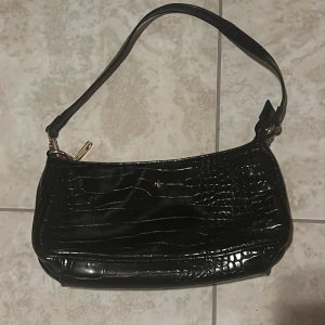 Svart axelväska i krokodilskinnsimitation  - Snygg svart axelväska med krokopräglat mönster och guldfärgade metalldetaljer. Köpt på TJmax. 