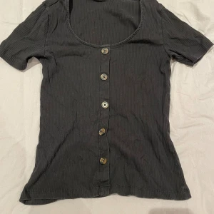 Svart ribbad topp med knappar - Svart ribbad topp från Gina Tricot med rund halsringning och fem knappar framtill. Kortärmad och figurnära passform. Perfekt att styla med jeans eller kjol för en chill look.