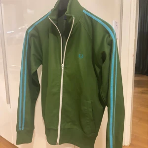 Grön Fred Perry hoodie med blå ränder - Snygg grön hoodie från Fred Perry med blå ränder längs ärmarna och vit dragkedja framtill. Klassisk sportig look med ribbade muddar och Fred Perry-logga på bröstet. Perfekt för dig som gillar retrostil och streetwear. Strl som herr x-Small till Small.