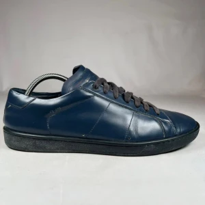 Mörkblå sneakers från Saint Laurent - Snygga mörkblå sneakers från Saint Laurent i slät skinn med svarta snören och svart sula. Klassisk låg modell med rund tå och diskret logga i guld på plösen. Perfekta för dig som gillar stilrena och exklusiva skor med minimalistisk vibe.