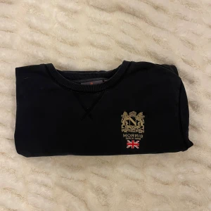 Svart sweatshirt från Morris - Svart sweatshirt från Morris med guldfärgat broderat emblem och engelsk flagga på bröstet. Tröjan har rund hals, ribbade muddar och lång ärm. Materialet är mjuk bomull som är skönt mot huden. Perfekt för dig som gillar klassisk stil med en twist. Värde 2000kr