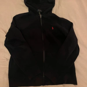 Svart hoodie från Polo Ralph Lauren - Svart hoodie från Polo Ralph Lauren med dragkedja och huva. Klassisk röd logga broderad på bröstet och känguruficka framtill. Tillverkad i mjuk bomull, perfekt för chill dagar. Långärmad och stilren design som funkar året runt.