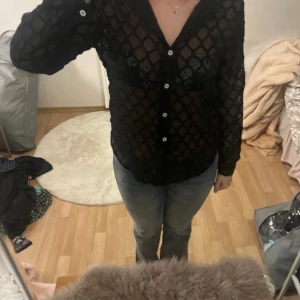 Svart transparent skjorta med knappar - Säljer en svart transparent skjorta med rutigt mönster och vita knappar. Skjortan har lång ärm och är i ett lätt, luftig material som ger en cool look. Perfekt för dig som vill sticka ut med en edgy stil.