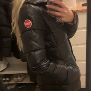 Svart dunjacka från Colmar - Snygg svart dunjacka från Colmar med quiltad design och logga på ärmen. Jackan har en figurnära passform, dragkedja och knappar framtill samt beige insida. Perfekt för kyliga dagar. Fått i present och har tyvärr inget kvitto. Kan ej namnet på modellen då det är en gammal modell som inte säljs längre. Storlek 38 men passar mig som brukar ha xs. Har ett litet hål (se sista bilden) men inget som man lägger märke till. 