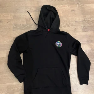 Svart Santa Cruz hoodie med färggrafik - Svart hoodie från Santa Cruz med stor färgglad grafisk print på ryggen och mindre tryck på bröstet. Tröjan har huva med snörning och en klassisk känguruficka framtill. Perfekt för dig som gillar streetwear och vill sticka ut med coola detaljer.  Tröjan säljs för 800+ nypris. Säljer för 150 kr nästan oanvänd.