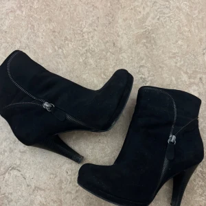 Svarta stövletter med klack - Säljer ett par svarta stövletter i mocka-look med markerad klack och rund tå. Skorna har en snygg dragkedjedetalj på sidan som ger en cool touch. Perfekta för dig som vill ha en stilren men edgy look till höst och vinter.