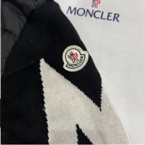 Svart Moncler cardigan  - Säljer en svart pufferjacka från Moncler med huva och snörning. Jackan har en dragkedja framtill och en unik vit zigzag-detalj på ena ärmen samt Moncler-logga. Materialet är en mix av polyester och stickad textil, perfekt för dig som vill ha en snygg och varm jacka.