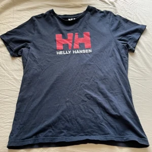 Svart Helly Hansen t-shirt med röd logga - Snygg svart t-shirt från Helly Hansen i storlek M. Framtill finns en stor röd HH-logga och vit text. T-shirten har rund hals och korta ärmar, tillverkad i mjuk bomull. Perfekt för dig som gillar sportiga och stilrena plagg med tydlig branding.