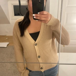 Beige ribbad kofta  - Snygg beige kofta från 157 i storlek M. Modellen är ribbstickad med v-ringning och tre knappar framtill. Croppad passform som sitter perfekt till jeans eller kjol. Långärmad och tillverkad i mjuk bomull för skön känsla. Perfekt för lager-på-lager under hösten.