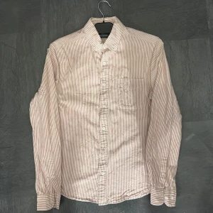 Randig skjorta från Dressmann, slim fit - Säljer en snygg randig skjorta från Dressmann i slim fit-modell. Skjortan är vit med tunna röda ränder, har klassisk krage, bröstficka och vita knappar. Perfekt för dig som gillar stilrena och tidlösa plagg.