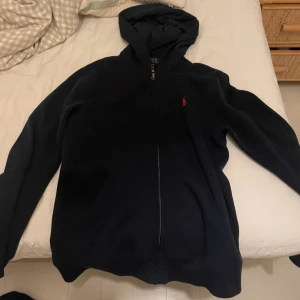 Svart hoodie från Polo Ralph Lauren - Svart hoodie från Polo Ralph Lauren med röd broderad logga på bröstet. Tröjan har huva, dragkedja framtill och två fickor. Perfekt för chill dagar och har en klassisk, enkel look som funkar till allt.