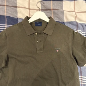 Olivgrön GANT pikétröja i bomull - Snygg olivgrön pikétröja från GANT med klassisk krage och två knappar framtill. T-shirten har korta ärmar och GANT-logga broderad på bröstet. Tillverkad i mjuk bomull som känns skön mot huden. Perfekt för en chill och stilren look.