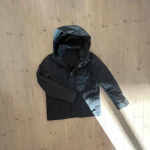 Svart parkajacka med huva - Svart parkajacka med stor huva och praktiska fickor framtill. Jackan har en stilren design med långa ärmar och knäppning framtill. Perfekt för kyliga dagar och enkel att matcha med olika outfits.