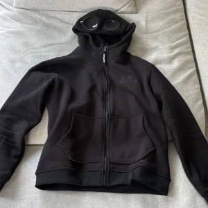 CP company hoodie - Svart hoodie från C.P. Company med ikoniska goggles i huvan och broderad logga på bröstet. Tröjan har dragkedja framtill, två stora fickor och är tillverkad i mjuk bomull. Perfekt för dig som vill sticka ut med streetwear-vibe.
