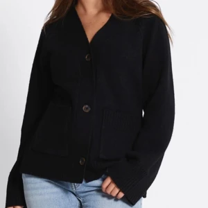 Svart kofta med knappar - Mysig svart kofta med stora knappar framtill och två rejäla fickor. Koftan har lång ärm och en avslappnad passform, perfekt att slänga på sig över en t-shirt. Ribbstickade detaljer vid ärmslut och fickor ger extra stilpoäng.