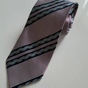  Yves Saint Laurent - Snyggt slips från Yves Saint Laurent med ett randigt mönster i marinblått, beige och blått samt små rutor i vinrött och vitt. Slipsen har en klassisk form och är tillverkat i siden, vilket ger en lyxig känsla. Perfekt för dig som vill sticka ut med en unik accessoar.